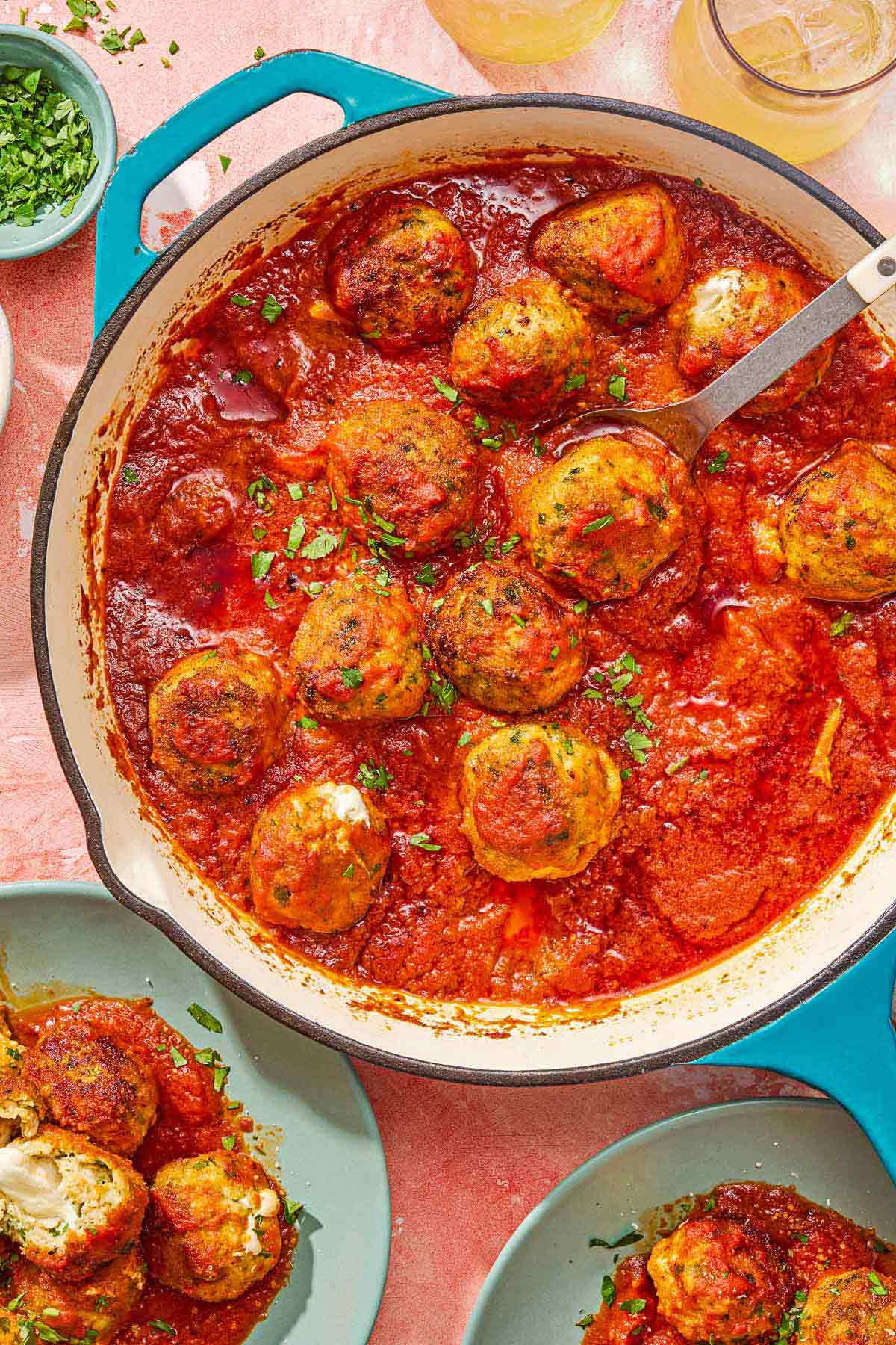 Mozzarella stuffed meatballs in a skillet with a serving spoon. Next to this is a bowl of chopped parsley, 2 beverages in glasses and 2 servings of the meatballs on plates.