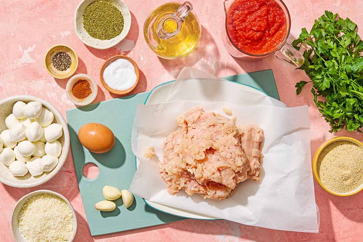 Ingredients for mozzarella stuffed meatballs including ground chicken, breadcrumbs, garlic, parsley, an egg, grated parmesan cheese, dried oregano, sweet paprika, salt, black pepper, baby mozzarella balls, olive oil and pasta sauce.