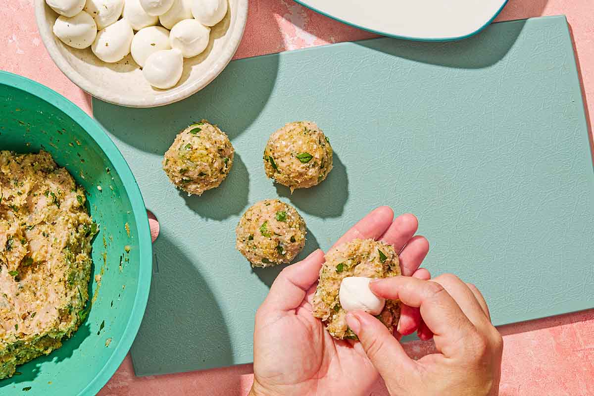 A raw meatball being stuffed with a baby mozzarella ball. In the background is a cutting board with 3 uncooked mozzarella stuffed meatballs, a bowl of the meatball mixture, and a bowl of baby mozzarella balls.