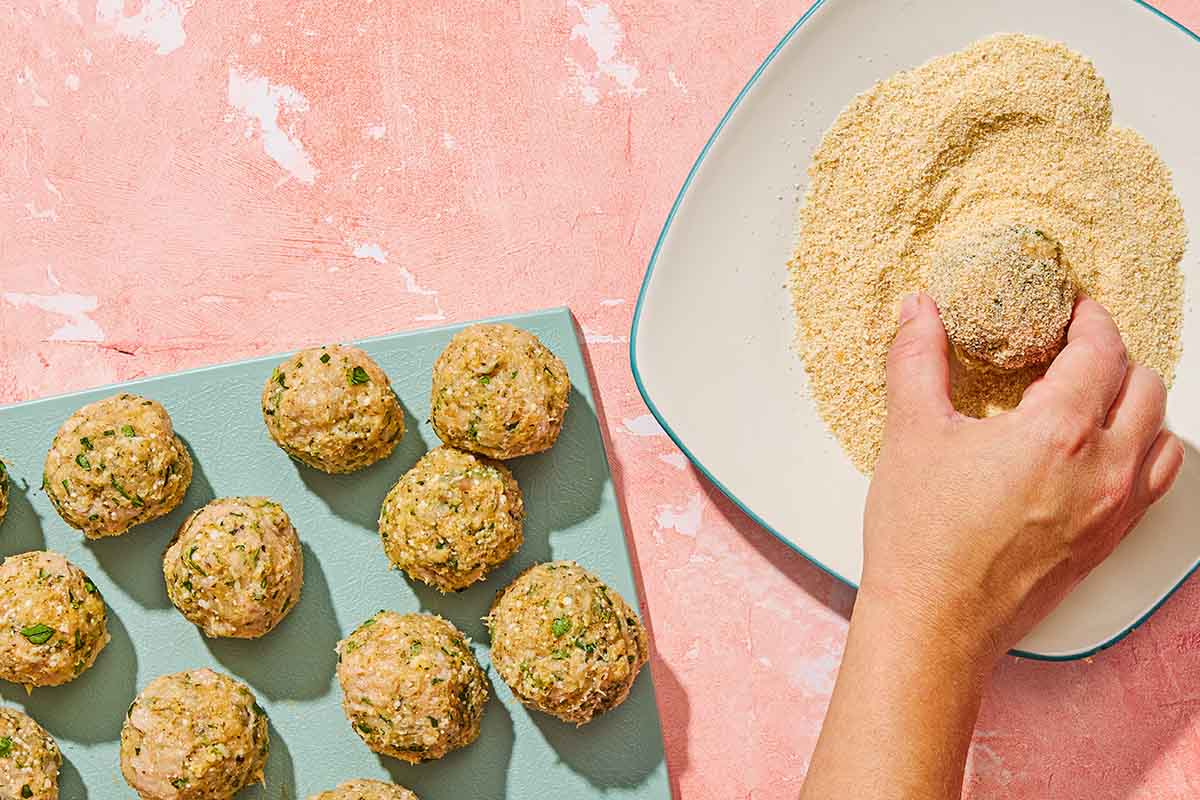 A cutting board with several uncooked, unbreaded mozzarella stuffed meatballs next to a plate where a meatball is being rolled in the breading.