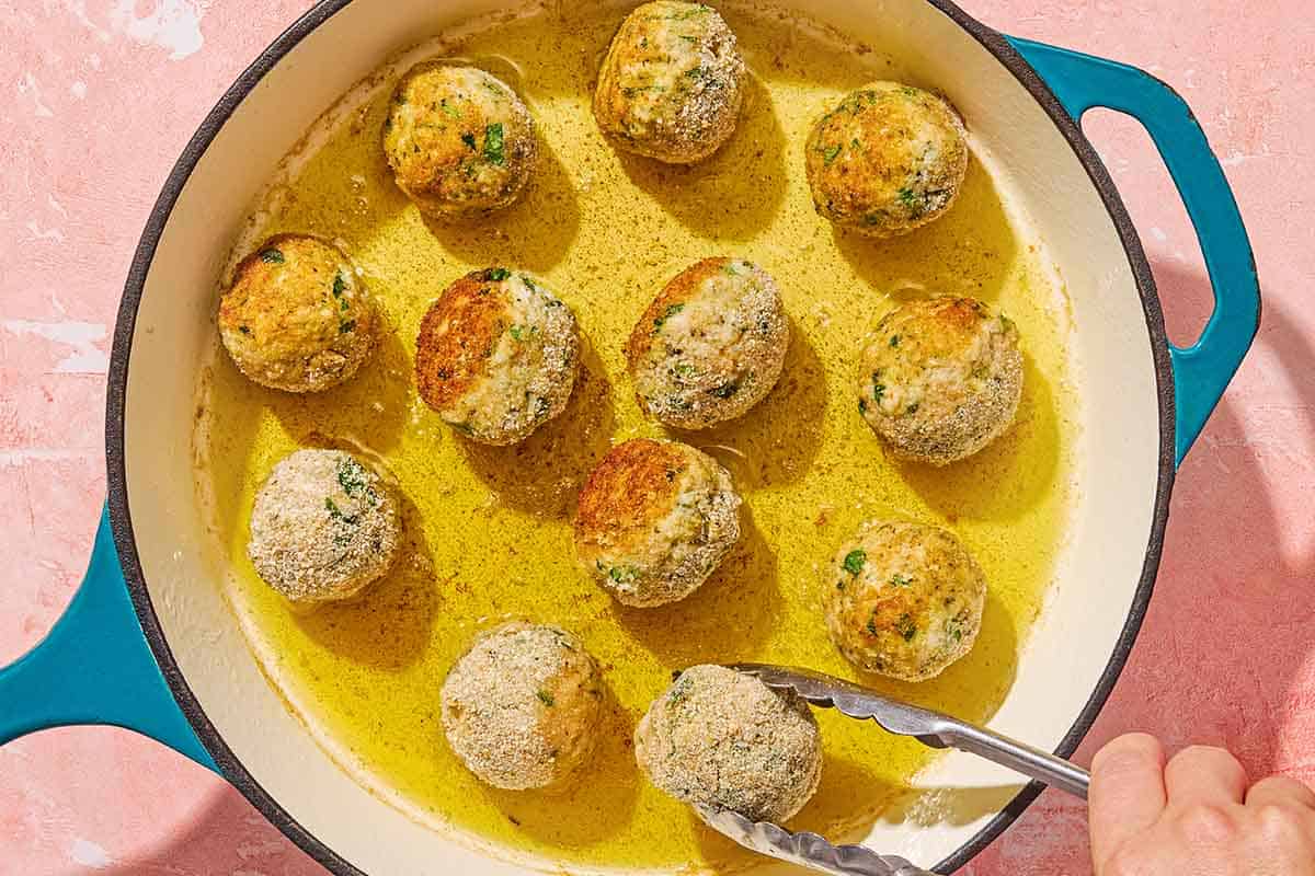 Mozzarella stuffed meatballs being fried in a skillet. One is being flipped with a pair of tongs.