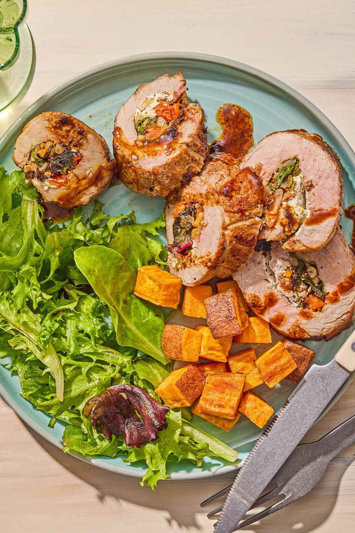A plate with several stuffed tenderloin pieces, salad and roasted sweet potatoes along with a knife and fork.