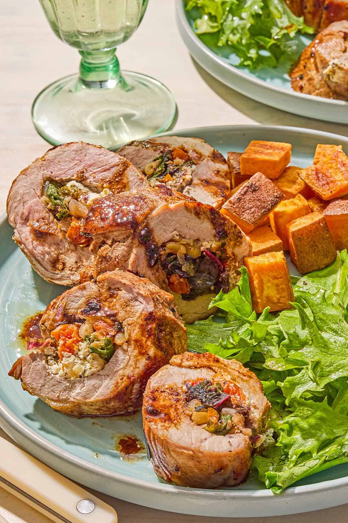 A close up of a plate with several stuffed tenderloin pieces, salad and roasted sweet potatoes. Next to this is another plate with the same meal and a glass of white wine.