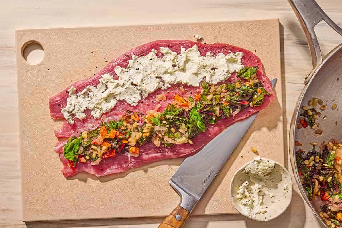 A butterflied pork tenderloin on a cutting board with a knife and a bowl of the goat cheese. On one half of the tenderloin is the goat cheese, and on the other half is the filling.