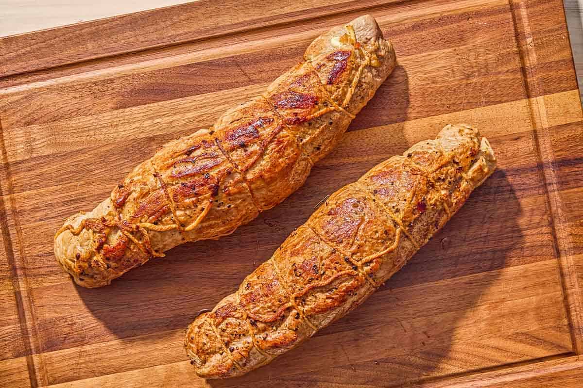 Two cooked, whole stuffed port tenderloins on a wooden cutting board.