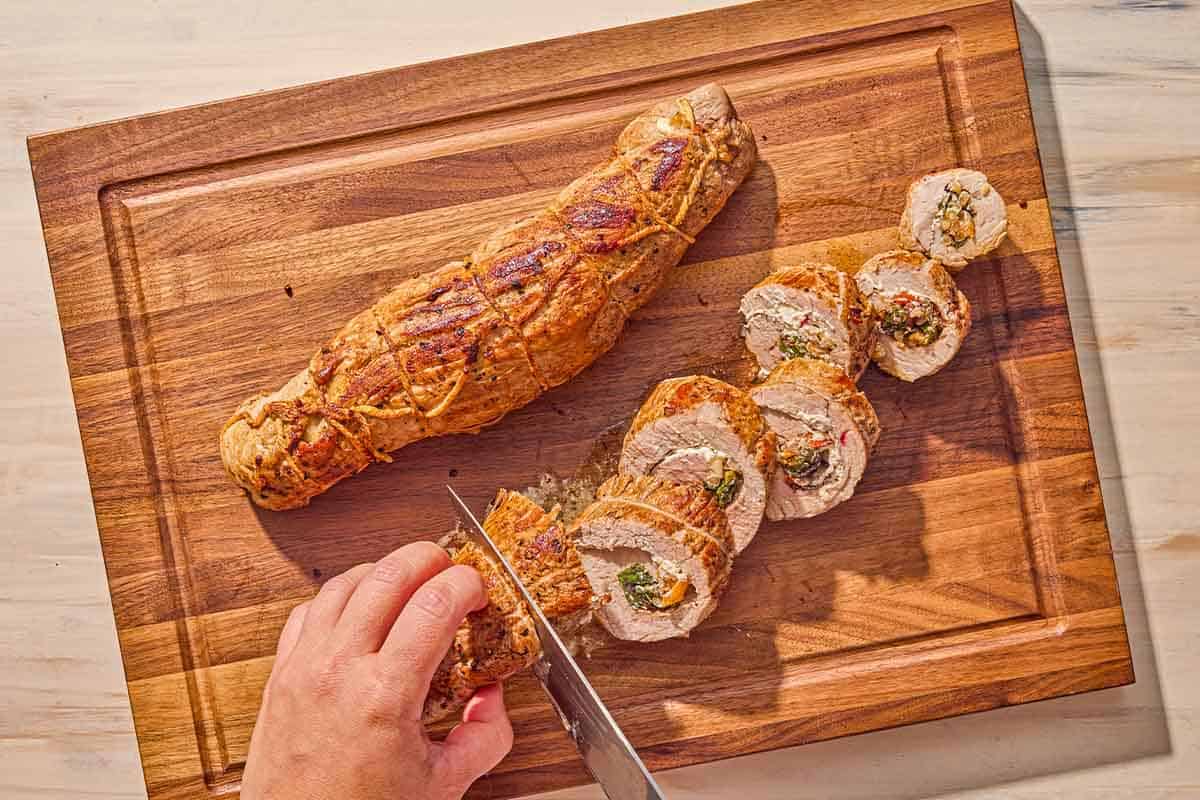 A cutting board with two stuffed pork tenderloins. One is being sliced into individual pieces with a knife.