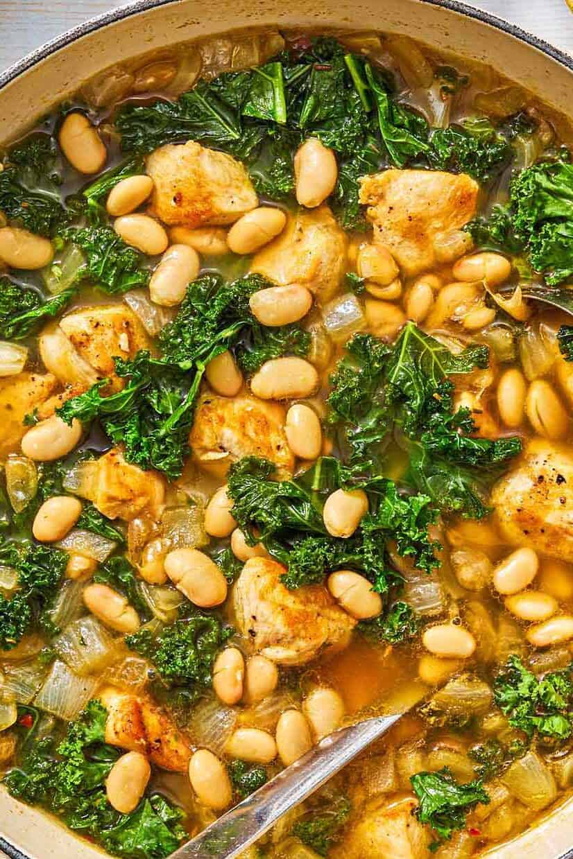 White bean and kale soup in a pot with a ladle next to a bottle oil and a lemon half.