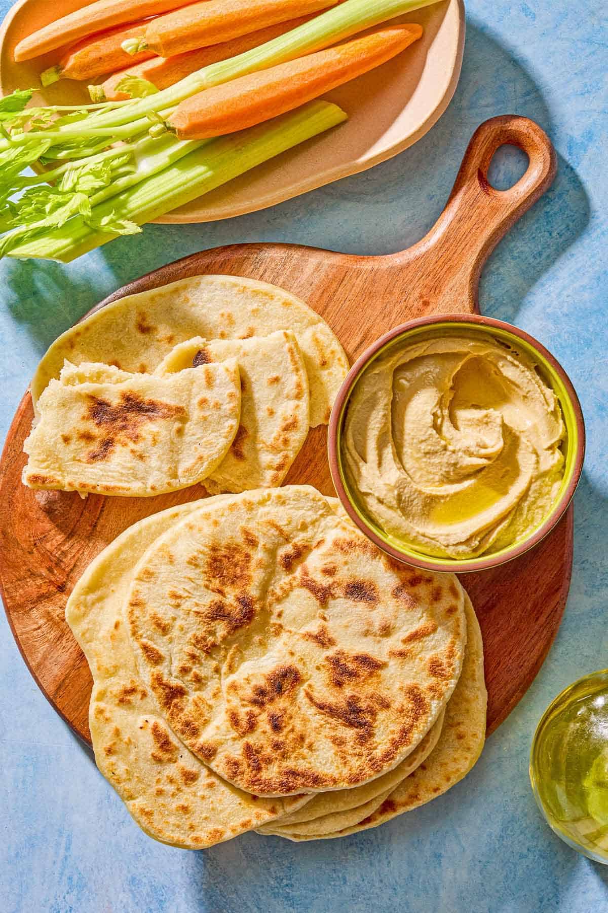 The pieces of the flatbread recipe on a wooden serving board with a bowl of hummus. Next to this is a plate with carrots and celery and a bottle of olive oil.