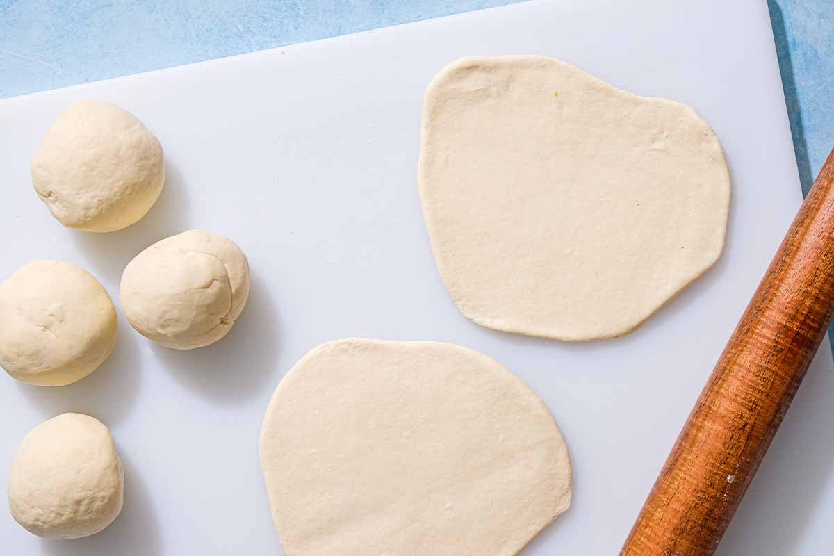 Four small balls of the dough for the flatbread recipe next to 2 rolled out disks of the dough along with a rolling pin.