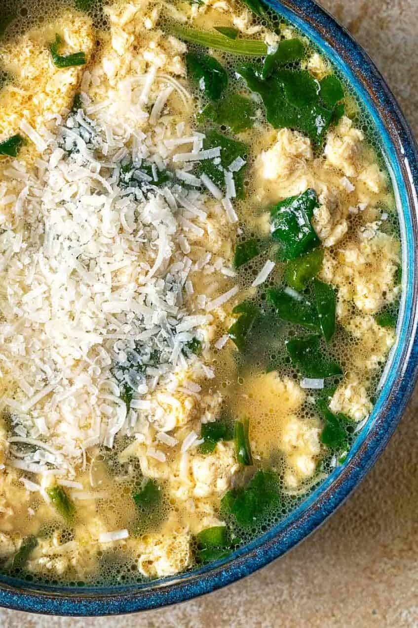 Overhead shot of a blue bowl with Stracciatella Soup with wilted spinach and freshly grated Parmesan cheese.