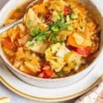 detox cabbage soup in a bowl with a spoon surrounded by small bowls of salt, pepper and lemon wedges and a cloth napkin.