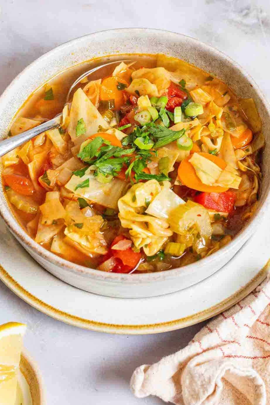 detox cabbage soup in a bowl with a spoon surrounded by small bowls of salt, pepper and lemon wedges and a cloth napkin.