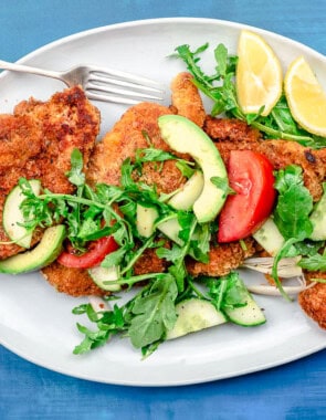 The chicken schnitzel recipe topped with arugula salad on a platter with a fork and lemon wedges.