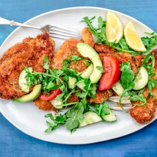 The chicken schnitzel recipe topped with arugula salad on a platter with a fork and lemon wedges.