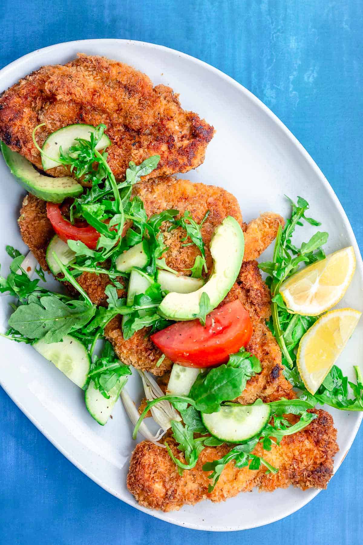 The chicken schnitzel recipe topped with arugula salad on a platter with lemon wedges.
