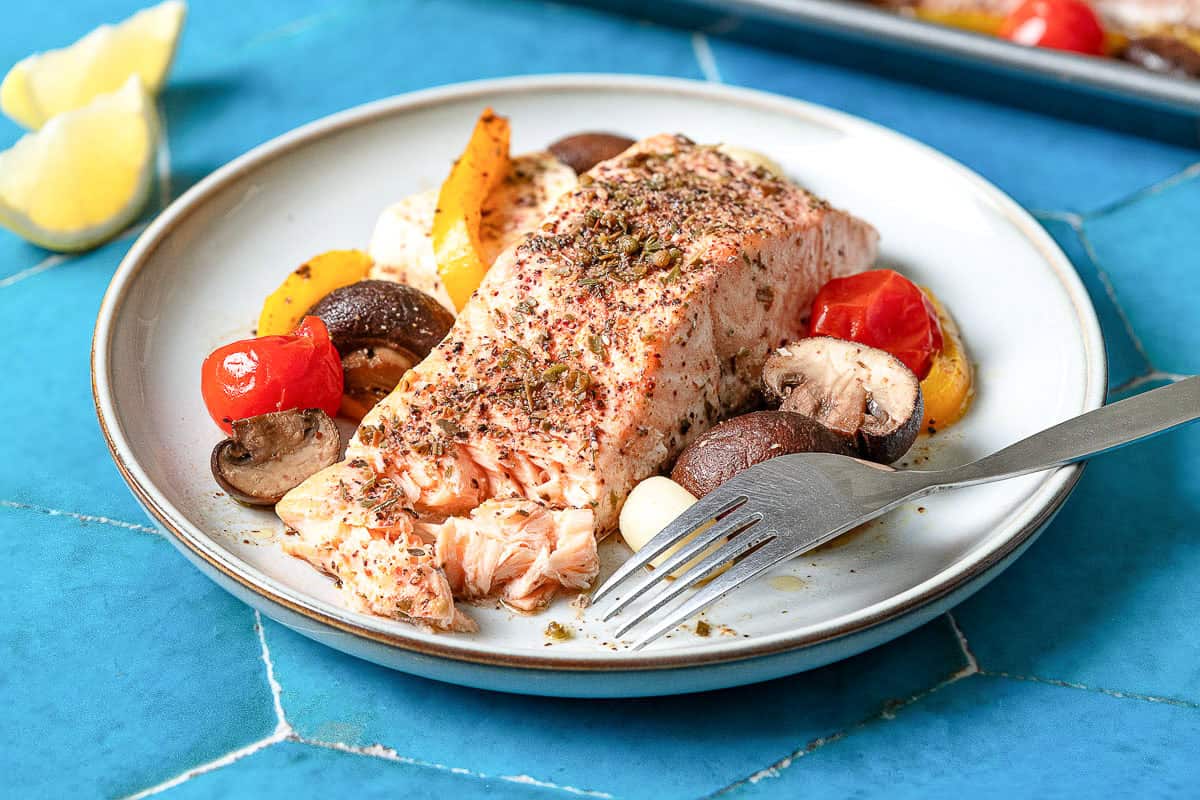 A serving of the mediterranean salmon recipe on a plate with a fork next to 2 lemon wedges.