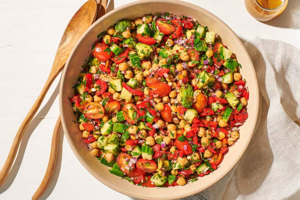 An overhead photo of the chickpea salad in a serving bowl next to a wooden serving spoon and fork, a jar of dijon dressing, and a cloth napkin.