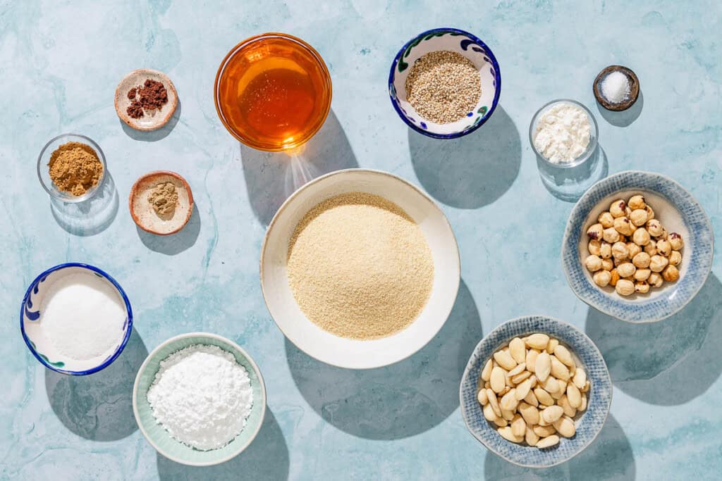 Ingredients for alfajores including blanched almonds, hazelnuts, sesame seeds, breadcrumbs, flour, cinnamon, coriander, cloves, salt, honey, powdered sugar and granulated sugar.