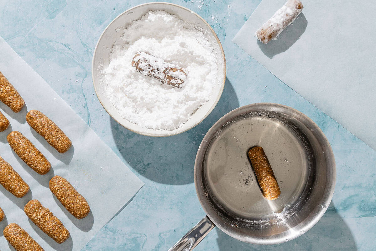 Uncoated alfajores spread evenly on a piece of parchment paper next to a saucepan with the simple syrup with on alfajores, a bowl of powdered sugar with one alfajores, and a finished alfajores on a piece of parchment paper.