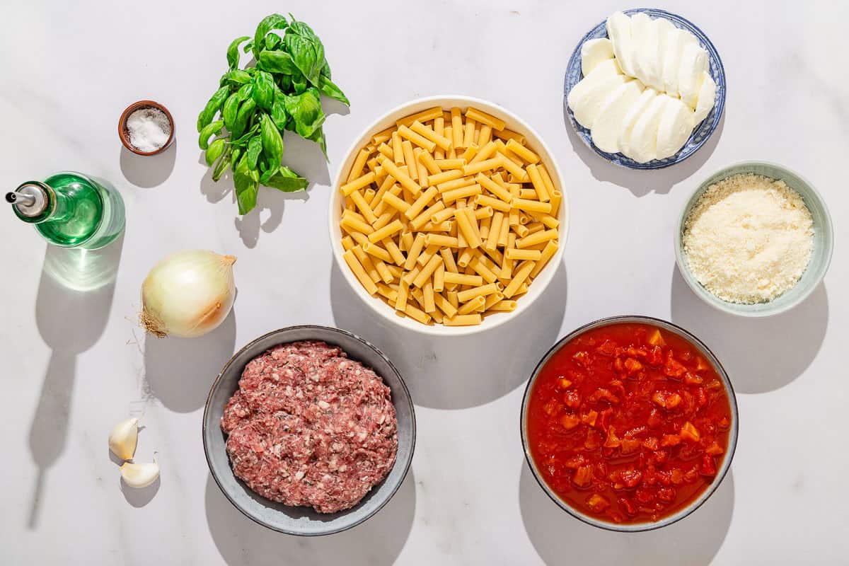 Ingredients for baked ziti including ziti, olive oil, salt, onion, garlic, Italian sausage, dice tomatoes, basil leaves, Parmesan cheese, and fresh mozzarella.