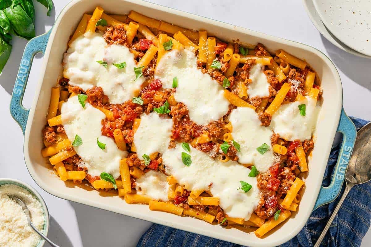 An overhead photo of baked ziti in a baking dish. Next to this is a serving spoon, a stack of two plates, basil leaves, and a bowl of grated Parmesan cheese.