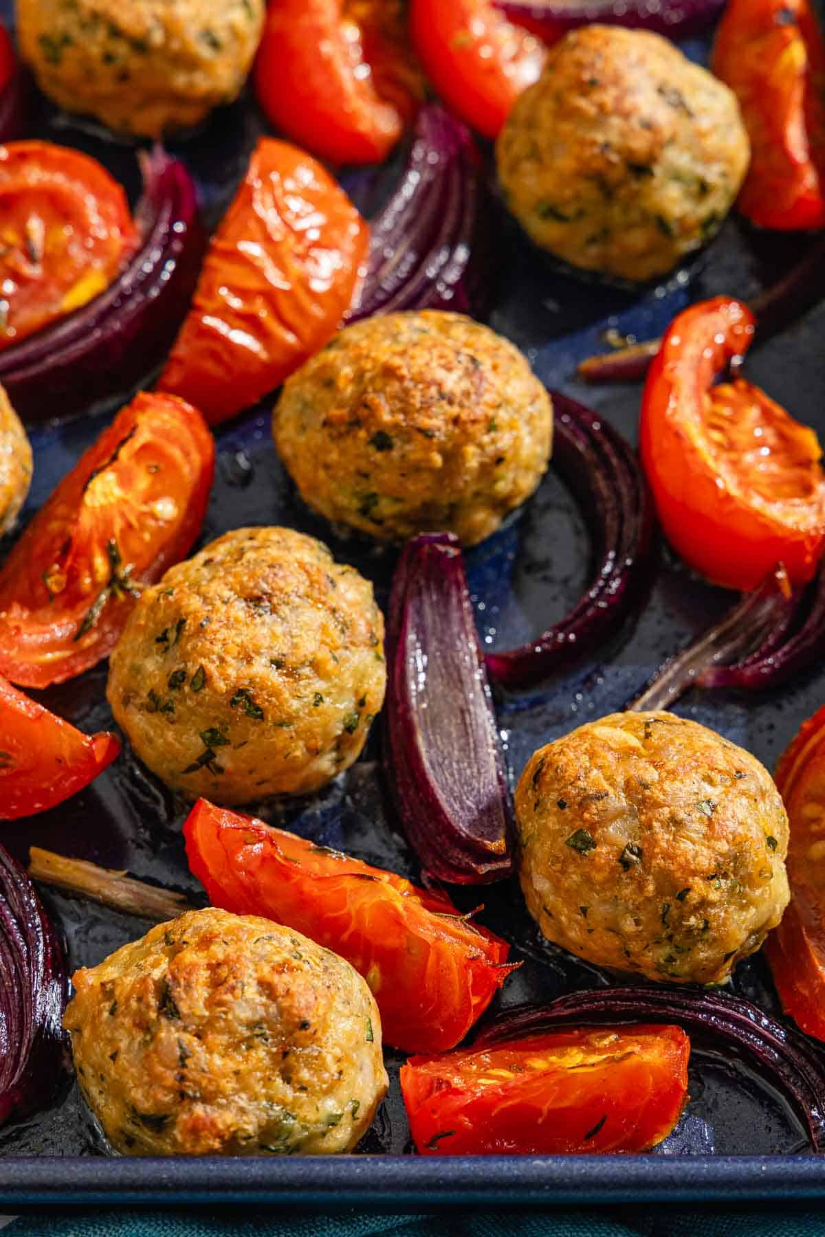 A close up of the baked chicken meatballs, tomatoes and red onions on a sheet pan.