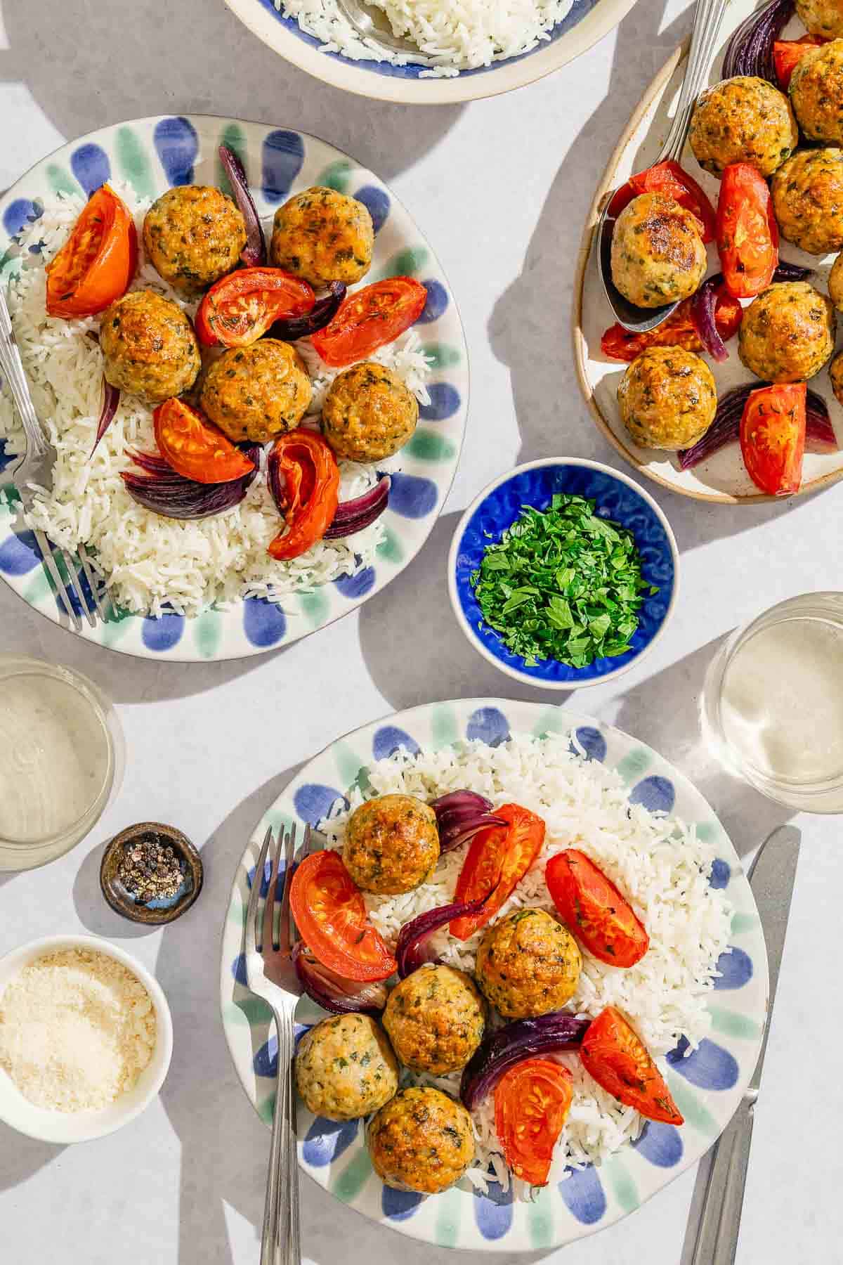An overhead photo of two servings of the baked chicken meatballs and rice on 2 plates with forks. Next to these are 2 glass of of white wine, small bowls of chopped parsley, grated parmesan and pepper, a knife, a bowl of rice, and the rest of the baked meatballs and veggies on a platter with a spoon.
