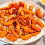 Honey glazed carrots topped with chopped parsley on a patter with a spoon. Next to this is a kitchen towel and bowls of cumin seeds, honey, and chopped parsley.
