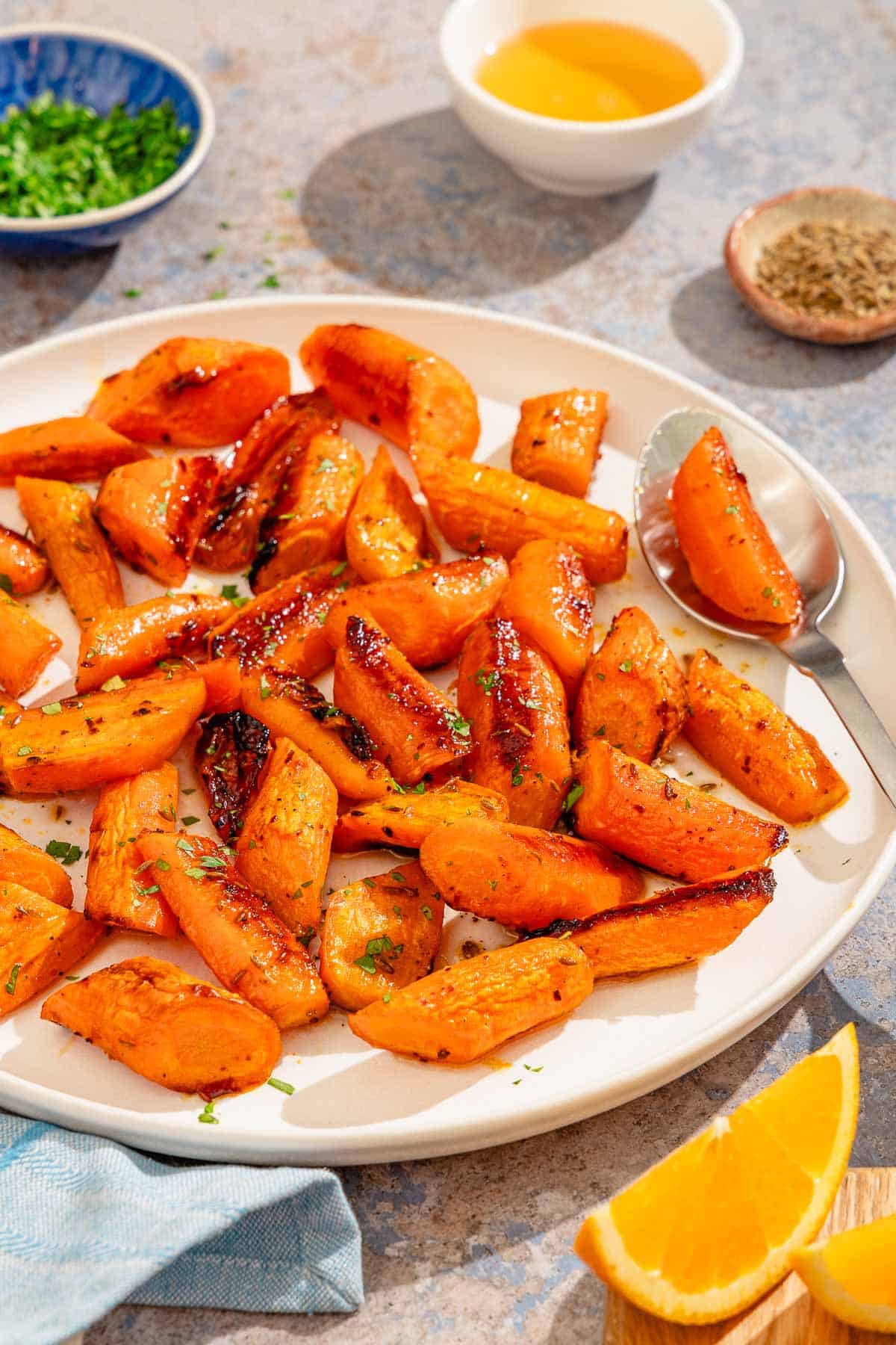 Honey glazed carrots topped with chopped parsley on a patter with a spoon. Next to this is a kitchen towel, orange wedges, and bowls of cumin seeds, honey, and chopped parsley.