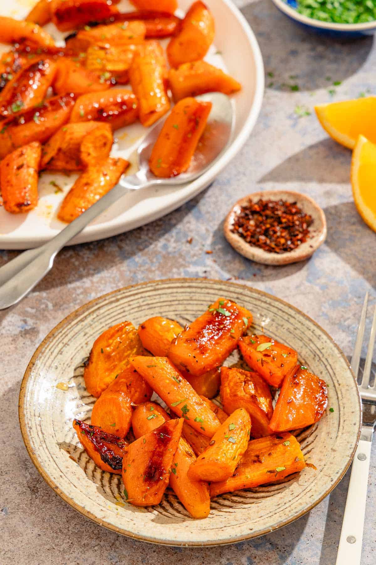 A serving of honey glazed carrots on a plate. Next to this is a fork, bowls of aleppo pepper and chopped parsley, orange wedges, and the rest of the carrots on a platter with a spoon.