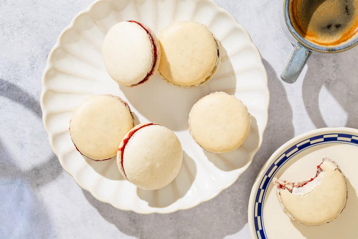Five macarons on a plate next to a plate with a single macron that has been partially eaten and a cup of coffee.