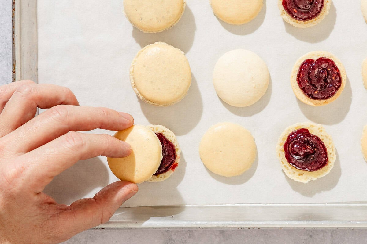 The top macaron shells being placed on top of the jam filled bottoms on a parchment lined sheet pan.