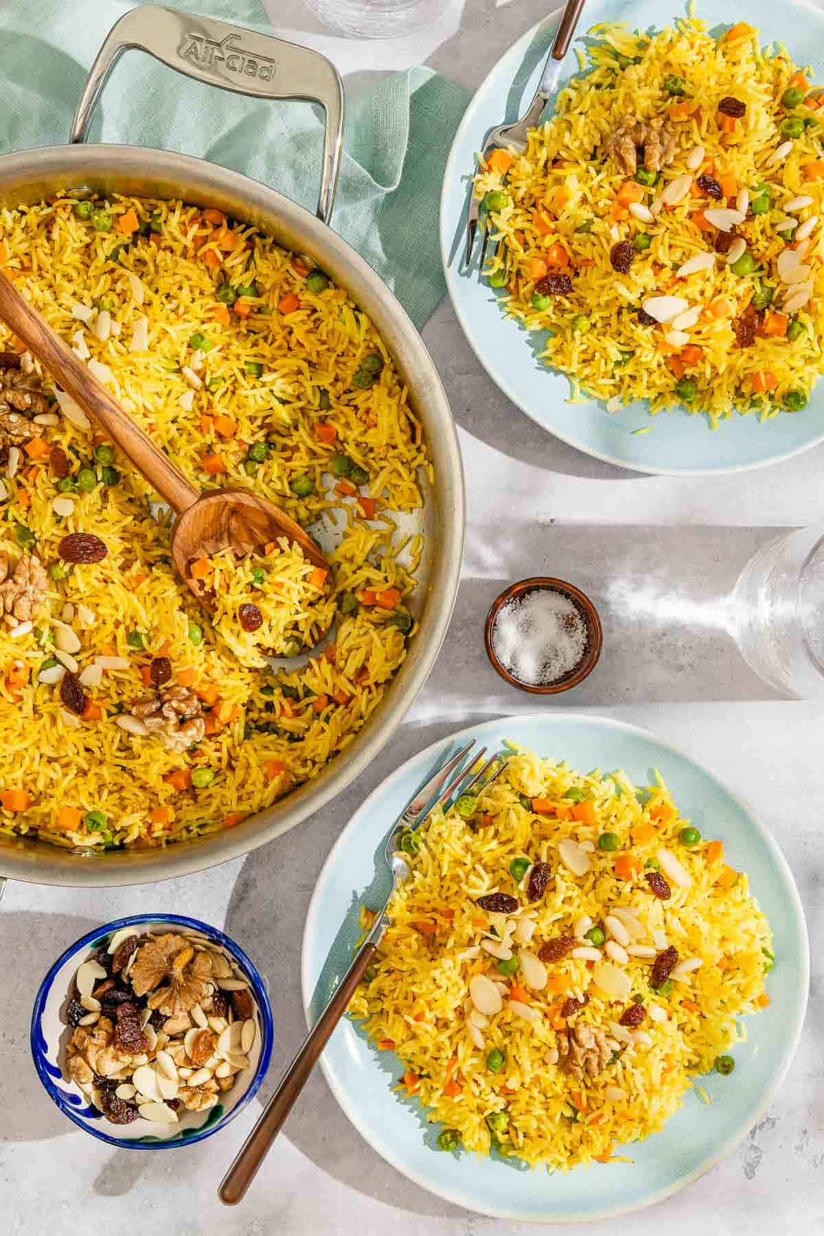 Two servings of the rice pilaf of plates with forks, Next to these a is the skillet with the rest of the rice pilaf recipe and a wooden spoon, a bowl of salt and a bowl of nuts and dried fruit.