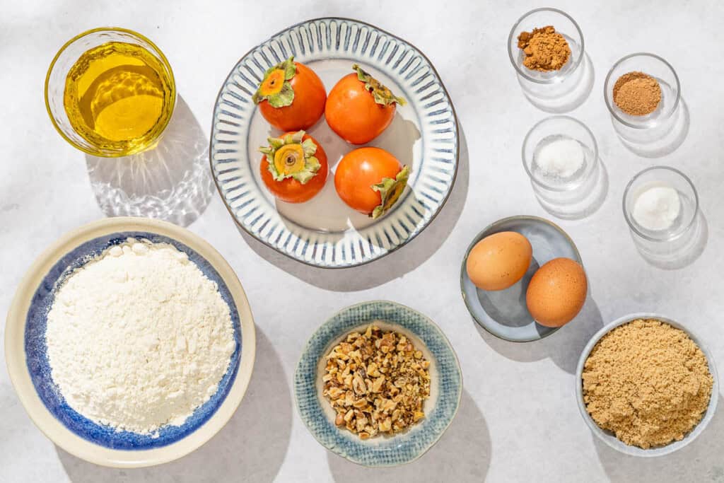 Ingredients fort the persimmon bread recipe including persimmons, brown sugar, olive oil, eggs, ground cinnamon, ground nutmeg, salt, flour, baking soda and toasted walnuts.