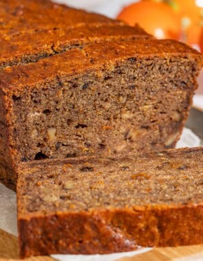 A partially sliced loaf of persimmon bread behind 3 slices of the bread on a parchment-lined cutting board with a knife. In the background are 3 persimmons.