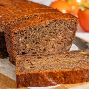 A partially sliced loaf of persimmon bread behind 3 slices of the bread on a parchment-lined cutting board with a knife. In the background are 3 persimmons.