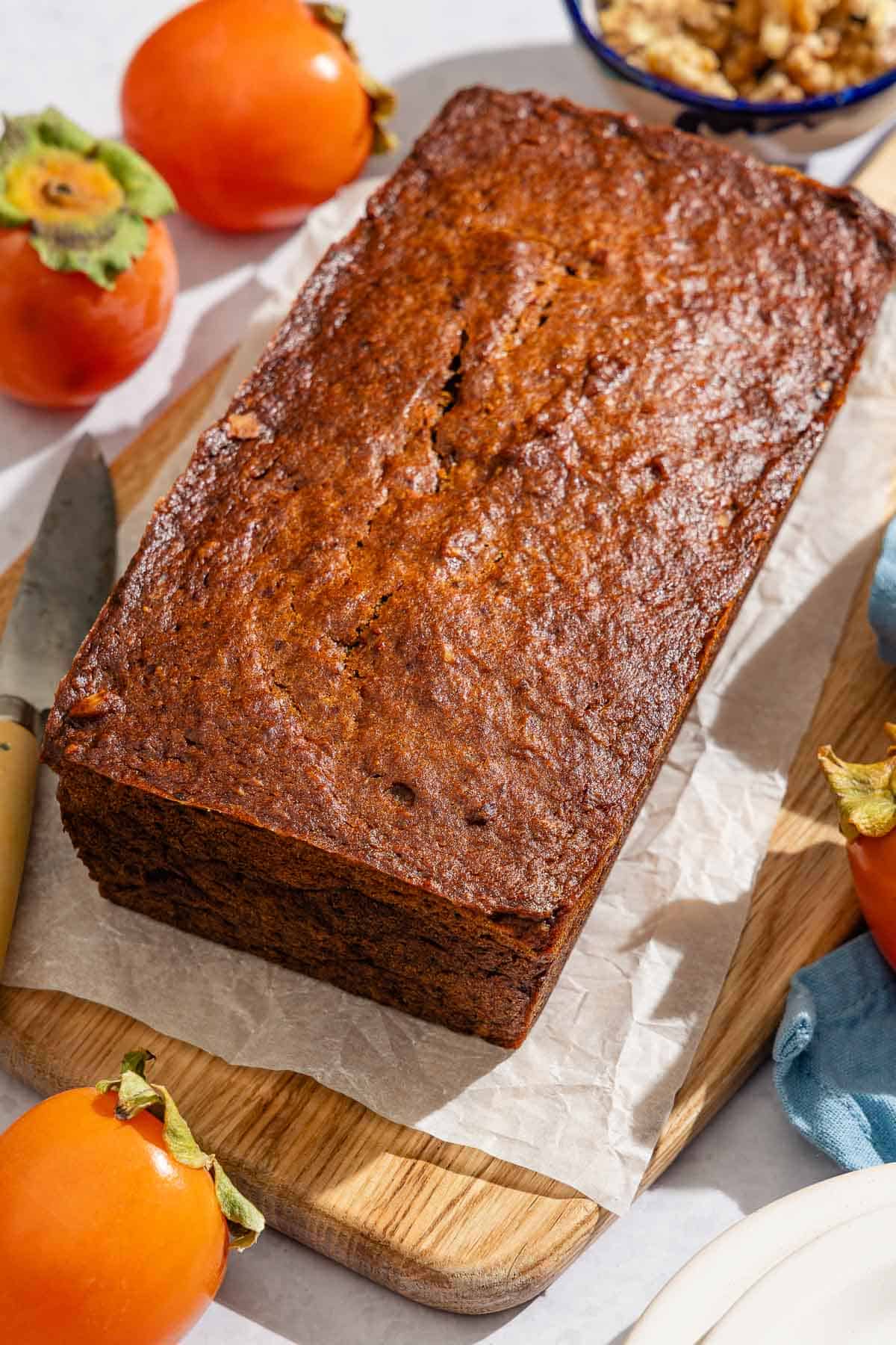 A loaf of persimmon bread on a parchment lined cutting board with a knife next to this is a bowl of walnuts and 3 persimmons.