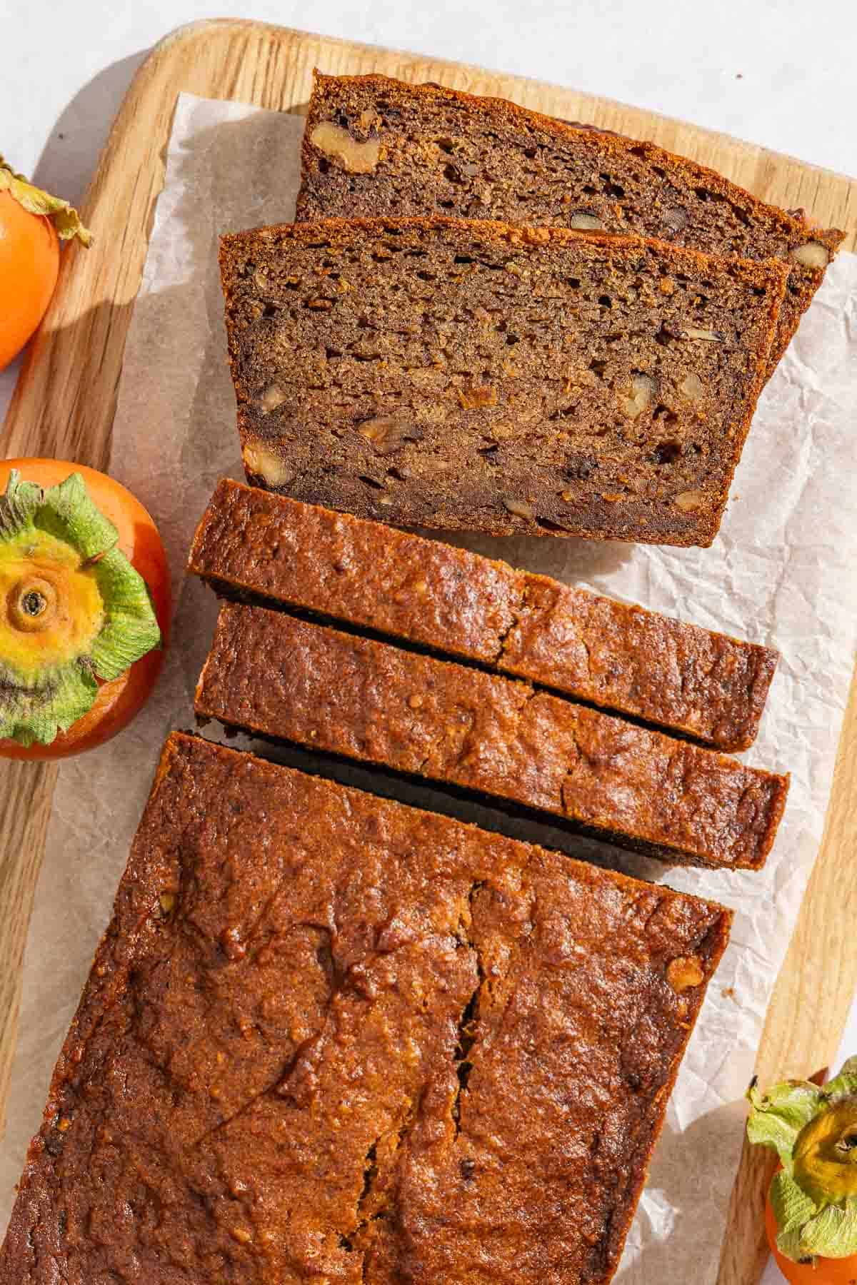 A partially sliced persimmon bread loaf next to 4 slices of the bread on a parchment lined cutting board. Next to this is three persimmons.