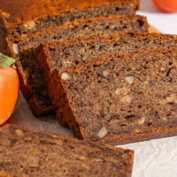 A partially sliced loaf of persimmon bread behind 3 slices of the bread on a parchment-lined cutting board with a knife. In the background are 3 persimmons.