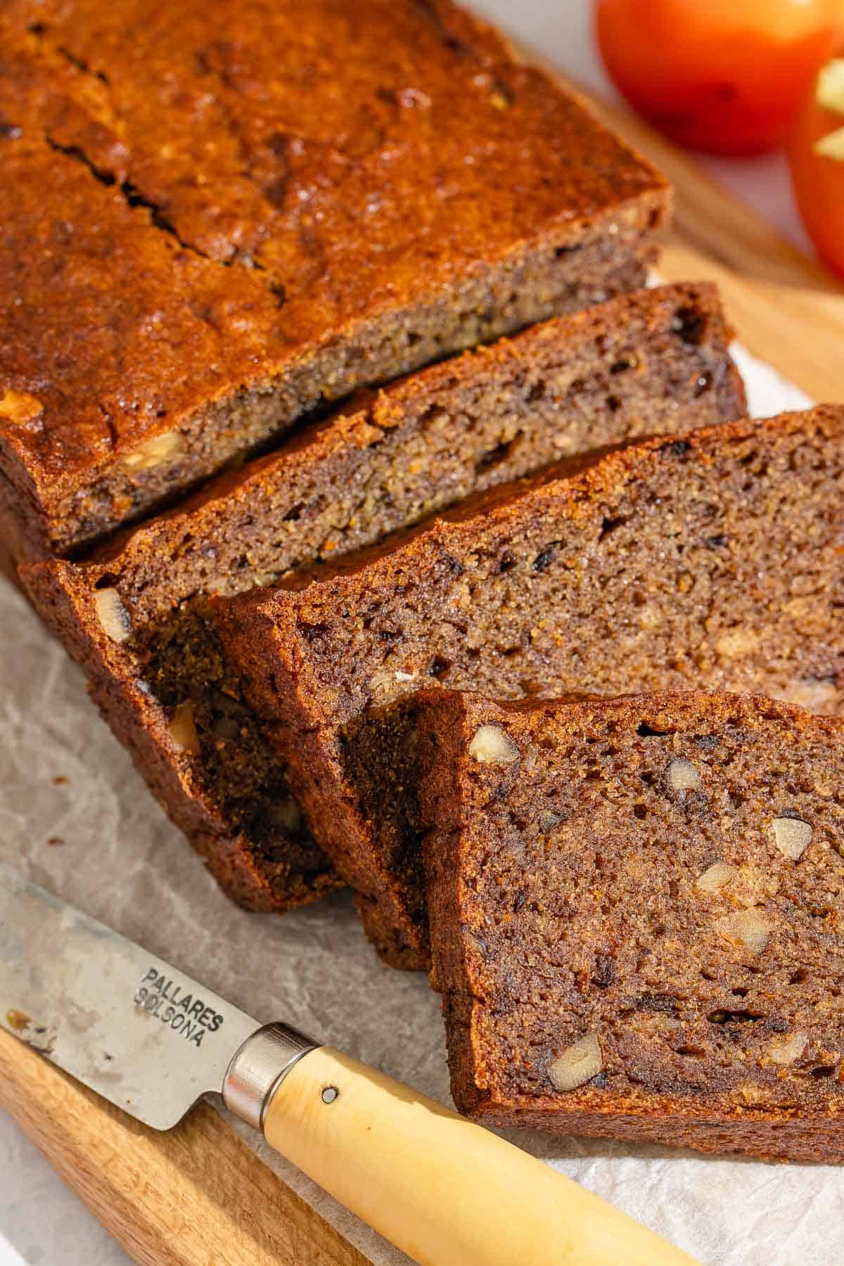 A partially sliced loaf of persimmon bread behind 3 slices of the bread on a parchment-lined cutting board with a knife.