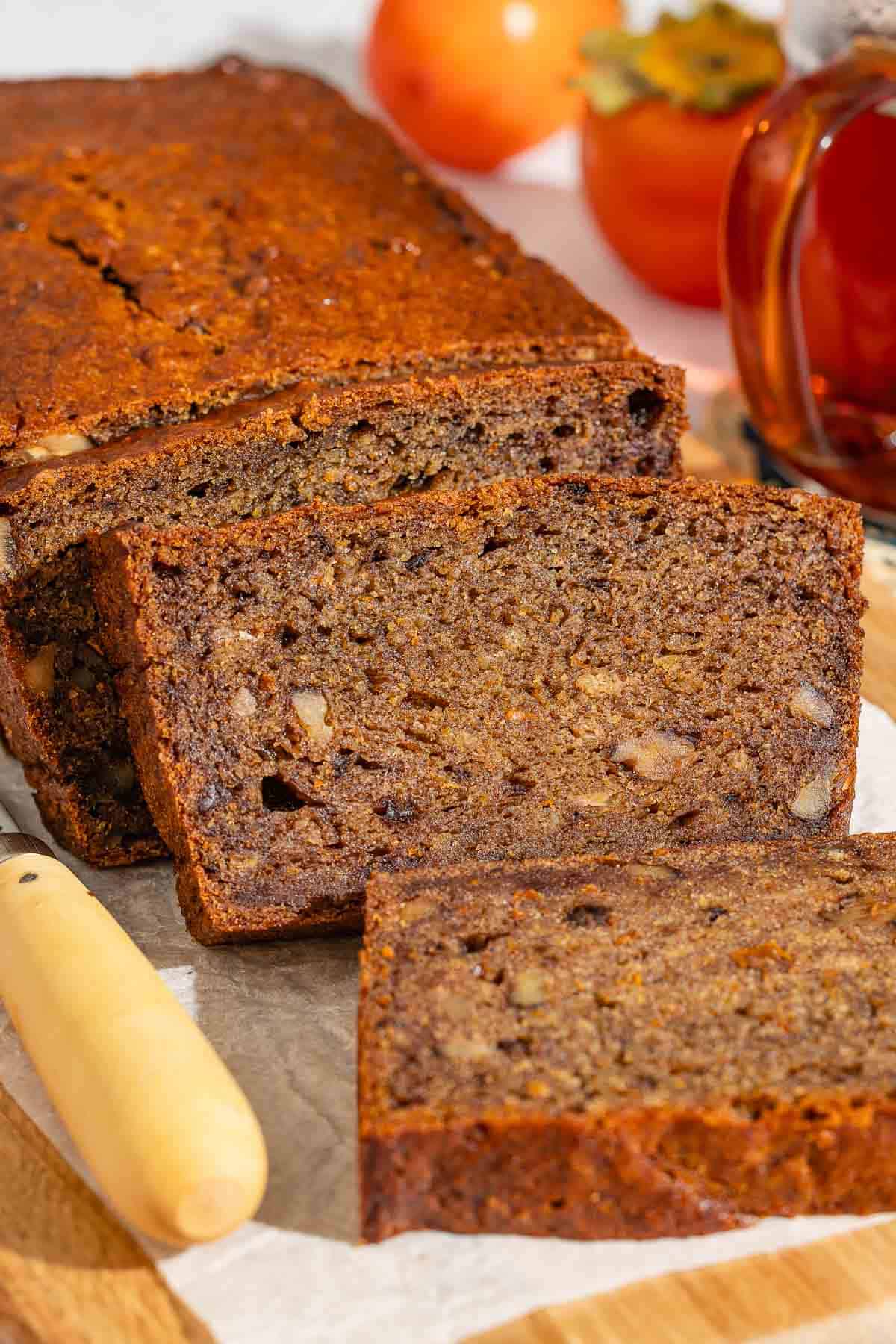 A partially sliced loaf of persimmon bread behind 3 slices of the bread on a parchment-lined cutting board with a knife. In the background are 3 persimmons and a cup of tea.