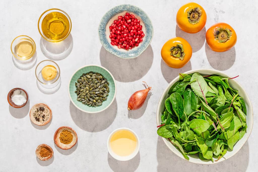 Ingredients for persimmon salad including persimmons, shallot, pepitas, mixed greens, pomegranate seeds, apple cider vinegar, honey, dijon mustard, cumin, cinnamon, salt, pepper, and olive oil.