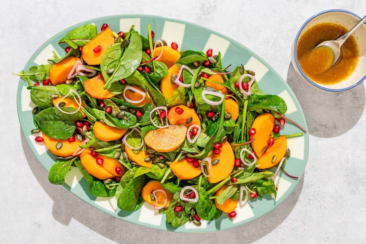 Persimmon salad on a platter next to a bowl of dressing with a spoon.