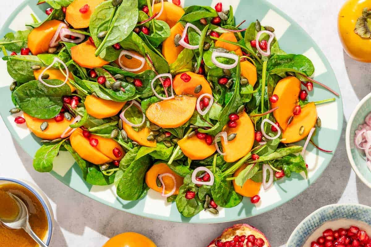 Persimmon salad on a platter surrounded by the dressing in a bowl with a spoon, a pomegranate, a bowl of pomegranate seeds, a bowl of sliced shallots, and a persimmon.