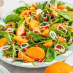 Persimmon salad on a platter next to the dressing in a bowl with a spoon, a stack of 2 plates, and 3 persimmons.