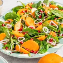 Persimmon salad on a platter next to the dressing in a bowl with a spoon, a stack of 2 plates, and 3 persimmons.