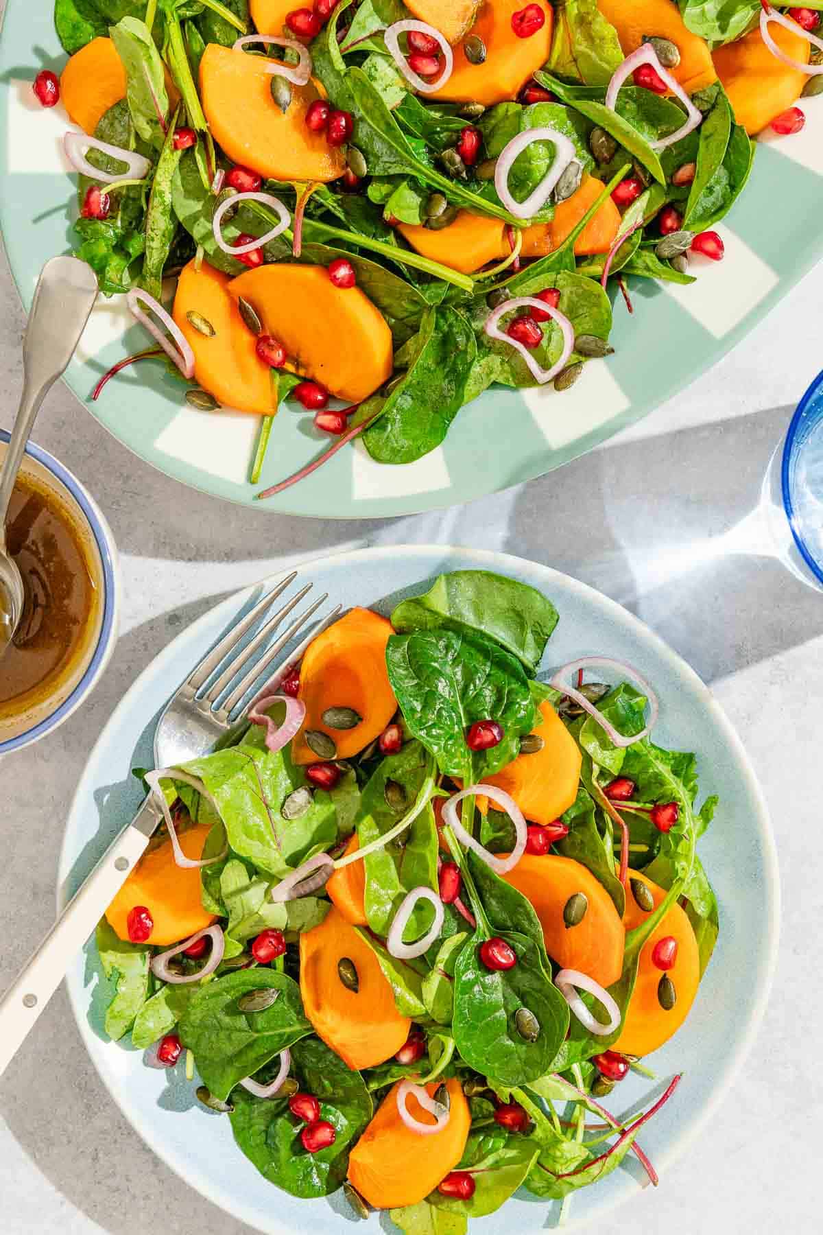 A serving of the persimmon salad on a plate with a fork. Next to this is the dressing in a bowl with a spoon, the rest of the salad on a platter, and a glass of sparkling water.