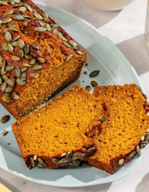 A partial pumpkin bread loaf along with 2 slices on a platter. Next to this is a knife, a bottle of ras el hanout, a bowl of ras el hanout, and a slice of pumpkin bread on a stack of 2 plates.