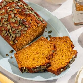 A partial pumpkin bread loaf along with 2 slices on a platter. Next to this is a knife, a bottle of ras el hanout, a bowl of ras el hanout, and a slice of pumpkin bread on a stack of 2 plates.