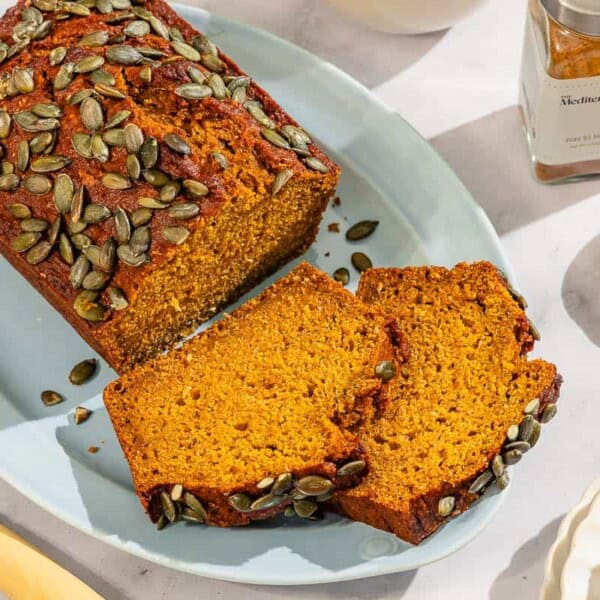 A partial pumpkin bread loaf along with 2 slices on a platter. Next to this is a knife, a bottle of ras el hanout, a bowl of ras el hanout, and a slice of pumpkin bread on a stack of 2 plates.